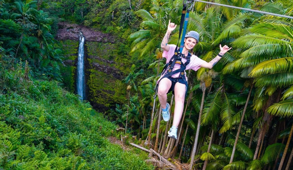 A smiling woman flashing a shaka sign while ziplining past a tropical waterfall surrounded by palm trees in Hawaii