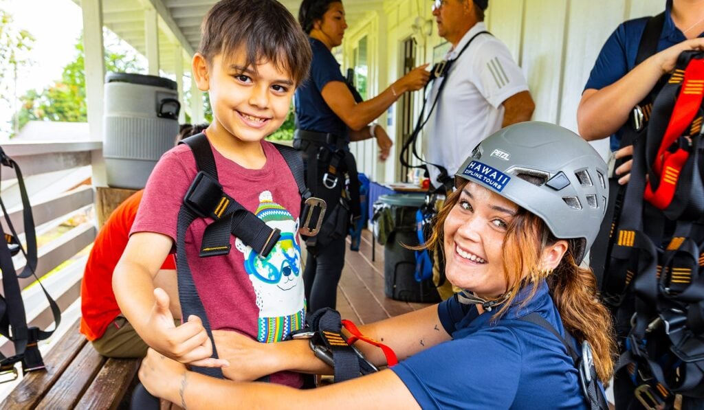 A Hawaii Zipline Tours guide in a Petzl helmet carefully fitting a safety harness on a young boy before his zipline ride
