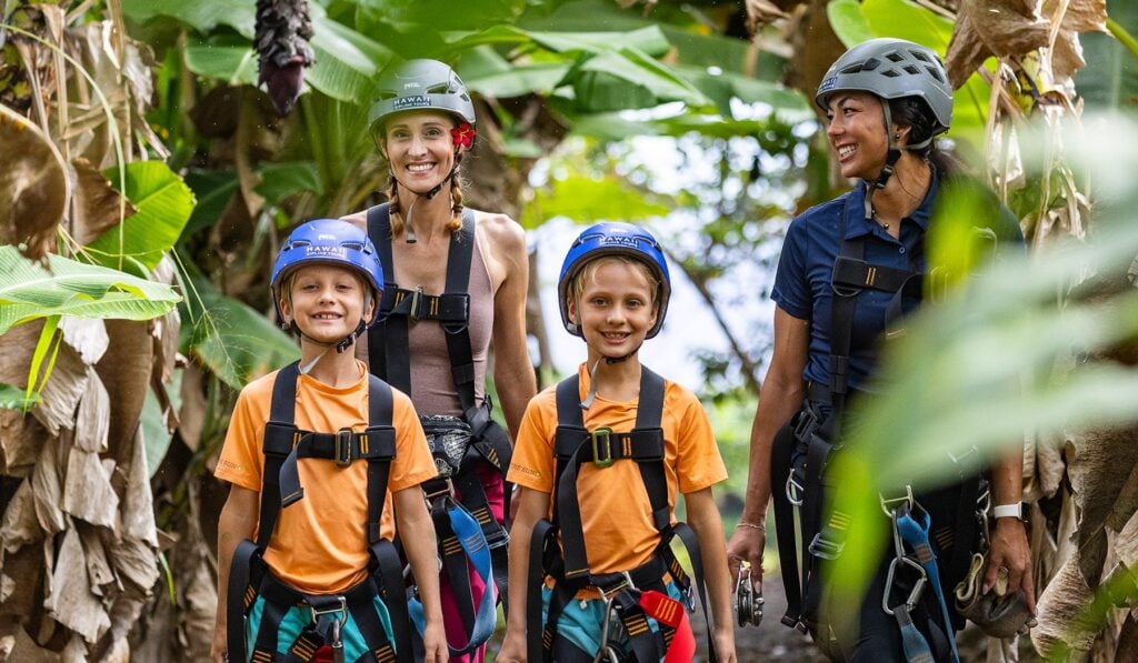 A family of four wearing helmets and harnesses smiles together on a Hawaii zipline tour amid lush tropical foliage