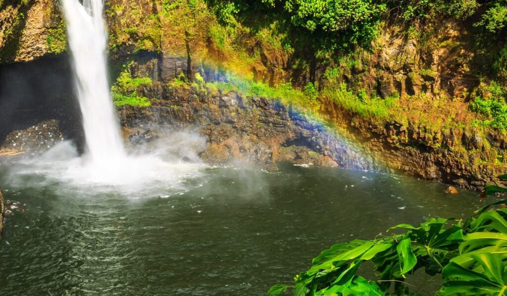 A rainbow forms in the mist at the base of a Hawaiian waterfall after rain, with vibrant green tropical vegetation in the foreground