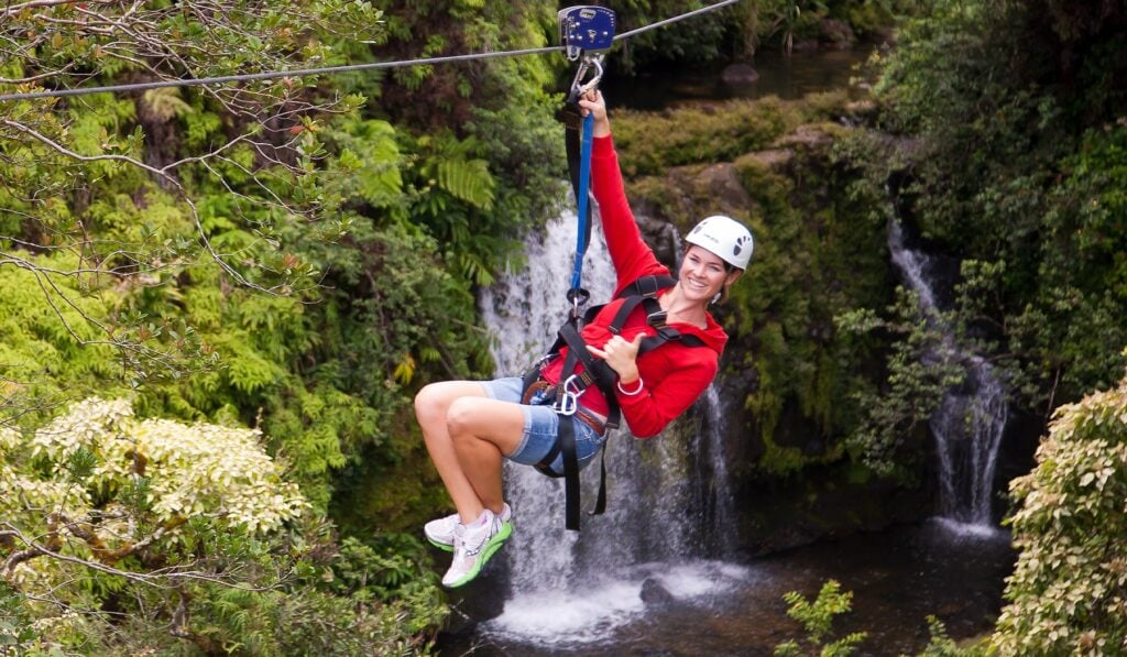 A woman in a red jacket gives a thumbs up while ziplining past a cascading Hawaiian waterfall, helmet and harness secured