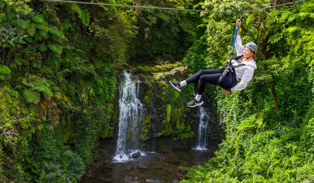 Guest ziplining in helmet and harness on a Hawaii Zipline Tours cable above a Big Island rainforest waterfall